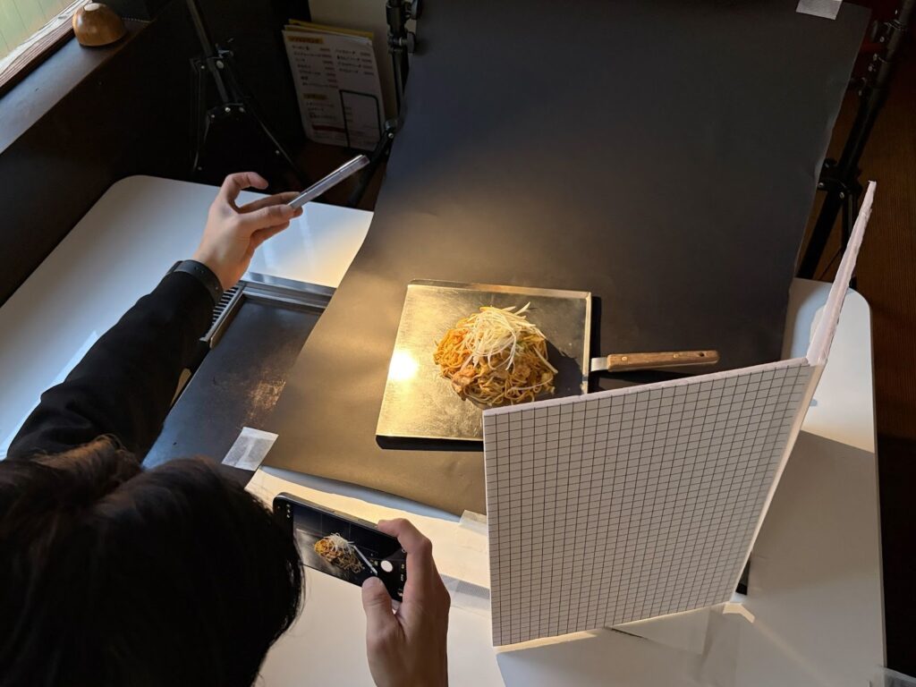 夜間営業の飲食店で別のスマホライトを斜め後ろから当てレフ板を挟んで撮影する様子